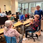 Kobieta siedzi przed komputerem, a mężczyzna pomaga jej w obsłudze urządzenia podczas warsztatów.