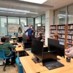 Mężczyzna prowadzi zajęcia w pracowni komputerowej z regałami pełnymi książek.