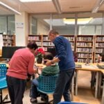 Mężczyzna pomaga dziecku w obsłudze urządzenia przy stole w otoczeniu regałów z książkami.