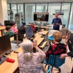 Uczestnicy warsztatów korzystają ze smartfonów podczas prezentacji w jasnym pomieszczeniu z dużymi oknami.