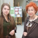 Dwie kobiety, jedna młodsza, druga starsza, pozują do zdjęcia obok planszy z informacjami o Powrocie Bractwa Śródziemia.