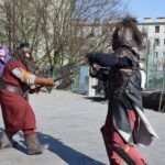 Dwóch rekonstruktorów w kostiumach postaci fantasy uczestniczy w inscenizacji walki na miejskim placu.