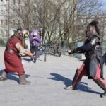Dwóch mężczyzn przebranych za wojowników z epoki fantasy trenuje szermierkę na placu miejskim.