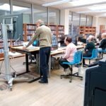 Uczestnicy warsztatów siedzą przy stolikach w bibliotece, otoczeni regałami z książkami.